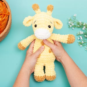 May include: A handmade, plush giraffe toy in shades of yellow and brown. The giraffe has a white muzzle, brown spots, and small, rounded ears. The toy is approximately 10 inches tall and is held against a light blue background.