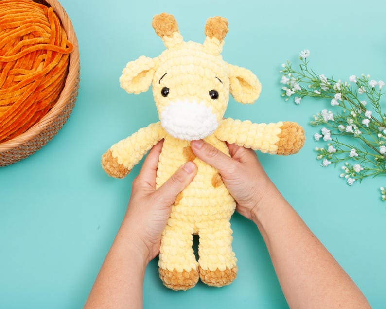 May include: A handmade, plush giraffe toy in shades of yellow and brown. The giraffe has a white muzzle, brown spots, and small, rounded ears. The toy is approximately 10 inches tall and is held against a light blue background.