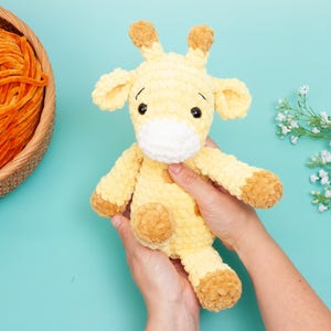 May include: A handmade, plush giraffe toy in shades of yellow and brown. The giraffe has a white muzzle, black eyes, and brown accents on its ears, hooves, and horns. A basket of orange yarn sits to the left. The toy is held against a light blue background.