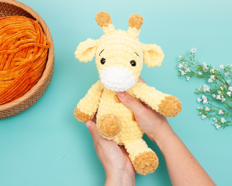 May include: A handmade, plush giraffe toy in shades of yellow and brown. The giraffe has a white muzzle, black eyes, and brown accents on its ears, hooves, and horns. A basket of orange yarn sits to the left. The toy is held against a light blue background.