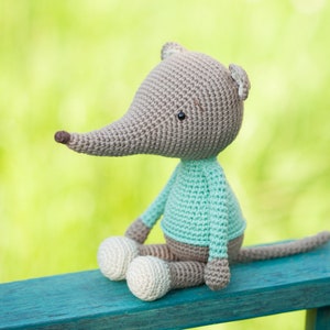 May include: A handmade crochet mouse doll with a long snout, small ears, and a long tail. The mouse wears a light blue sweater and has cream-colored feet. The doll is sitting on a blue surface against a blurred green background.