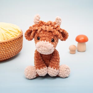 May include: A handmade brown and tan crochet cow plushie with small horns and a fluffy texture. The cow is sitting with its legs tucked in. A basket of yellow yarn and wooden mushrooms are in the background.