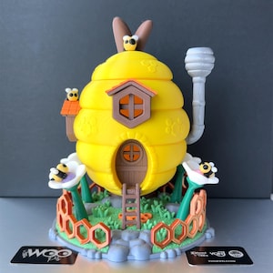Puede incluir: Una escultura caprichosa de colmena amarilla con techo, puerta y ventana marrones. La colmena está adornada con abejas de dibujos animados y se asienta sobre una base con flores y formas de panal. Una chimenea gris se extiende desde la parte superior. Es un objeto decorativo.