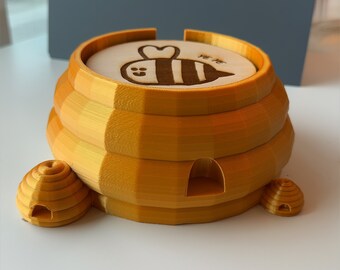 May include: A gold-colored 3D printed beehive-shaped coaster with a wooden top featuring a bee design and the text "Bee-lieve". The coaster has three small beehive-shaped feet.