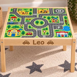 May include: A square wooden play table featuring a printed road map surface with buildings, trees, and vehicles. The front apron is engraved with the name Leo flanked by two car icons. The table has four light-coloured wooden legs and a flat top.