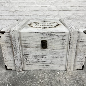 May include: A white wooden chest with a distressed finish, featuring metal corner brackets and a clasp. The top is adorned with a floral wreath and the words "Audrie & Evan Memories".