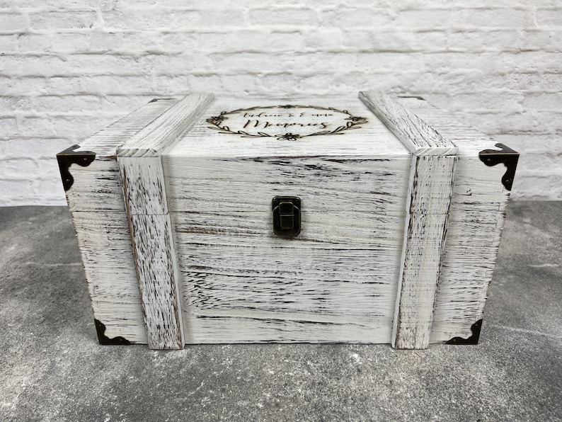 May include: A white wooden chest with a distressed finish, featuring metal corner brackets and a clasp. The top is adorned with a floral wreath and the words "Audrie & Evan Memories".