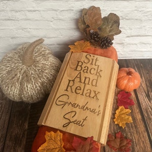 May include: A wooden sign with the text "Sit Back And Relax Grandma's Seat" engraved on it. The sign is surrounded by autumnal decorations, including pumpkins and leaves.