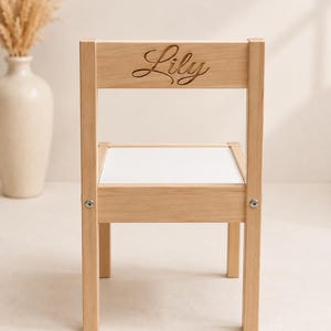 May include: A light-coloured wooden chair featuring a square seat with a white surface and a backrest engraved with the name Lily in a cursive script. The chair has four straight legs and visible metal hardware connecting the seat to the frame.