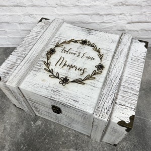 May include: A white wooden memory box with distressed paint and metal corner accents. The box features a decorative floral wreath with the names "Andrew & Emma" and the word "Memories" engraved on the lid.