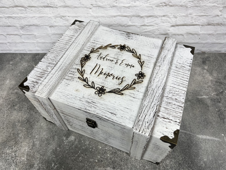 May include: A white wooden memory box with distressed paint and metal corner accents. The box features a decorative floral wreath with the names "Andrew & Emma" and the word "Memories" engraved on the lid.