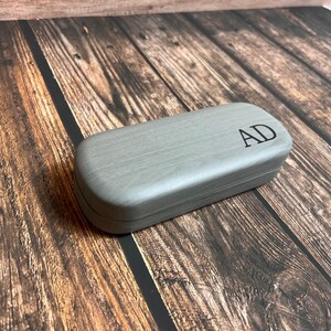 May include: A gray, rectangular glasses case with rounded edges. The case has a wood-grain texture and the initials "AD" are printed in black on the top. The case is closed and sits on a wooden surface.