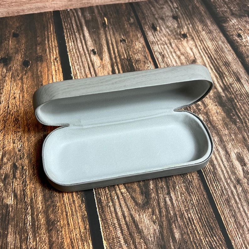 May include: An open, gray eyeglasses case with a wood grain pattern on the exterior. The interior is a light gray, soft material. The case is empty and ready to hold a pair of glasses.