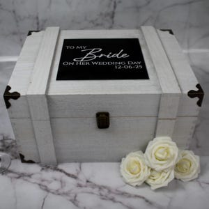 May include: A white wooden box with a hinged lid and metal hardware. The box has the text "TO MY Bride ON HER WEDDING DAY 12-06-25" on a black background. A small bouquet of white roses rests in front of the box.