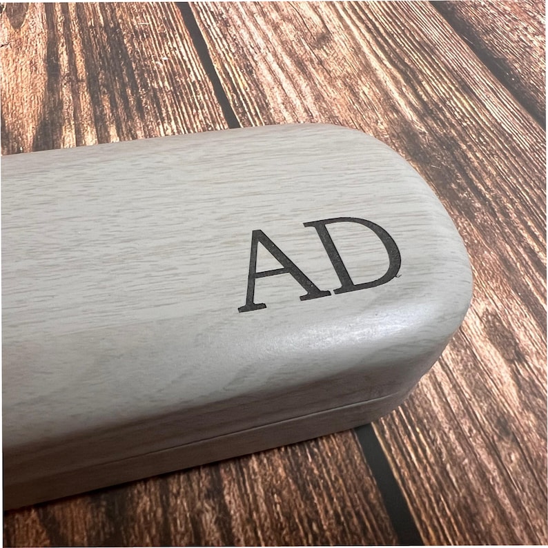 May include: A light gray wooden glasses case with the initials "AD" engraved in black. The case has a rectangular shape with rounded edges. The background is a wooden surface.