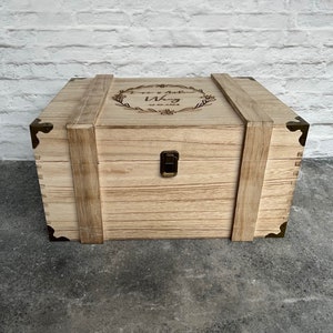 May include: A light-colored wooden box with metal corner accents and a clasp closure. The box has a hinged lid with a decorative wreath and text. The box is made of wood and has a rustic appearance.