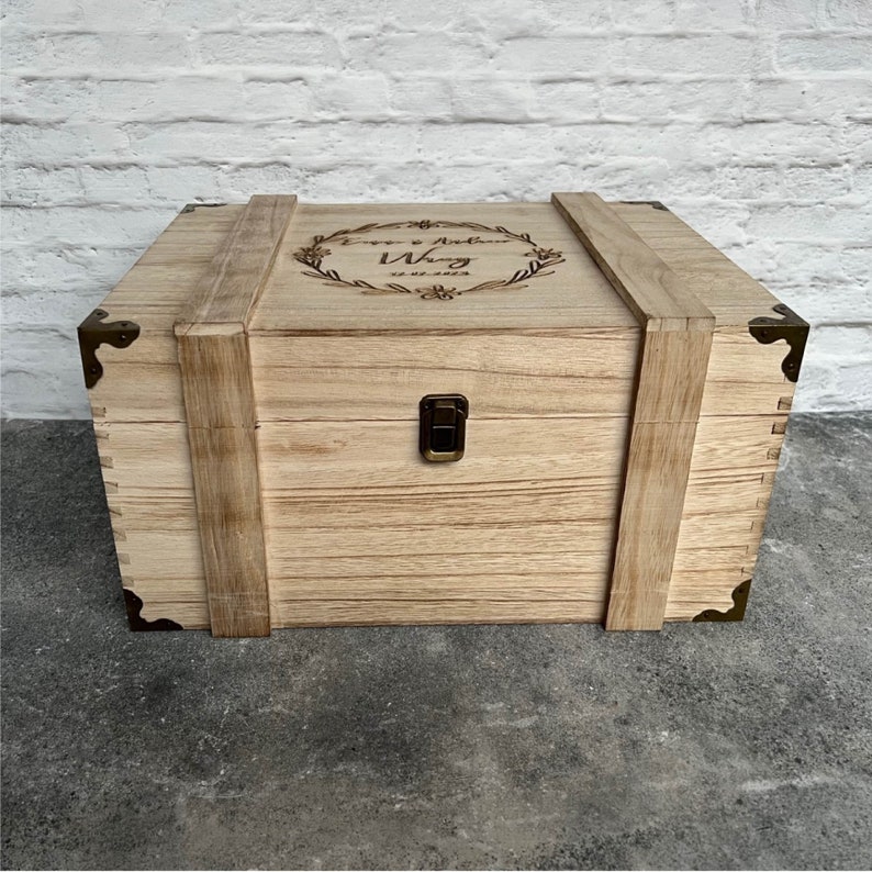 May include: A light-colored wooden box with metal corner accents and a clasp closure. The box has a hinged lid with a decorative wreath and text. The box is made of wood and has a rustic appearance.