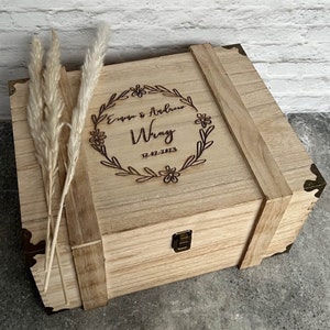 May include: A light-colored wooden box with a hinged lid, reinforced with wooden straps and metal corner brackets. The lid is engraved with the names "Evan & Andrew Wray" and the date "12-02-2023" within a floral wreath. Dried ornamental grass is placed on the left side.