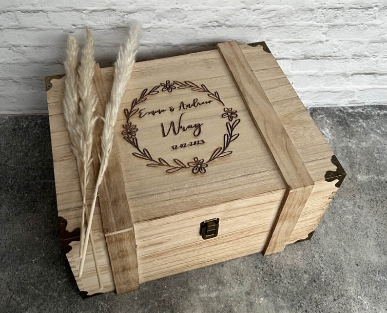 May include: A light-colored wooden box with a hinged lid, reinforced with wooden straps and metal corner brackets. The lid is engraved with the names "Evan & Andrew Wray" and the date "12-02-2023" within a floral wreath. Dried ornamental grass is placed on the left side.