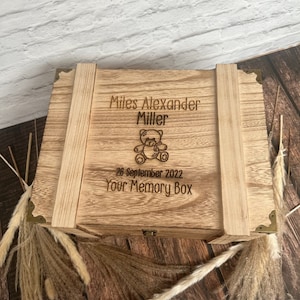 May include: A wooden memory box with a hinged lid and brass accents. The box is engraved with the name "Miles Alexander Miller", a teddy bear illustration, the date "26 September 2022", and the text "Your Memory Box".