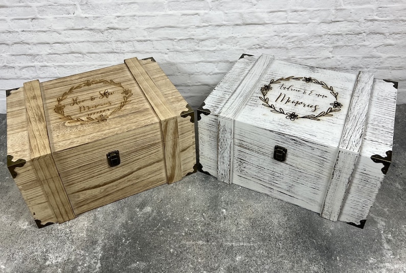 May include: Two wooden keepsake boxes, one light brown and one white, with decorative metal corner brackets and clasps. The light brown box has the text "Her & His Memories" in a floral wreath. The white box has the text "Adriana & Emma Memories" in a floral wreath.