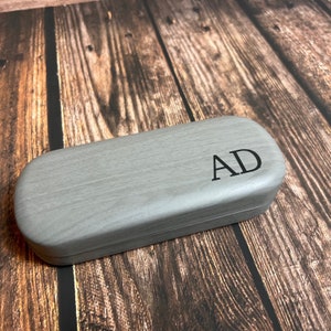 May include: A gray, rectangular glasses case with rounded edges. The initials "AD" are printed in black on the top. The case is made of a wood-like material and sits on a wooden surface.