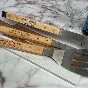 May include: A set of wooden-handled barbecue tools with stainless steel blades. The handles are engraved with phrases such as "Don't Worry, I Got This" and "Happy Father's Day." A beer bottle and wine glass are also in the image. The tools are on a wooden board.