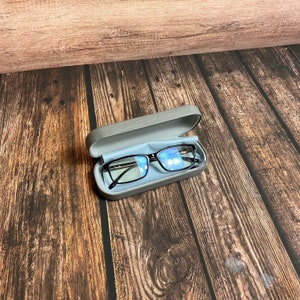 May include: Black-framed eyeglasses inside a gray eyeglasses case with a blue microfiber cloth. The case is open, revealing the glasses. The background is a wooden surface with a brown wood grain.