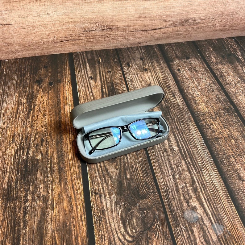 May include: Black-framed eyeglasses inside a gray eyeglasses case with a blue microfiber cloth. The case is open, revealing the glasses. The background is a wooden surface with a brown wood grain.