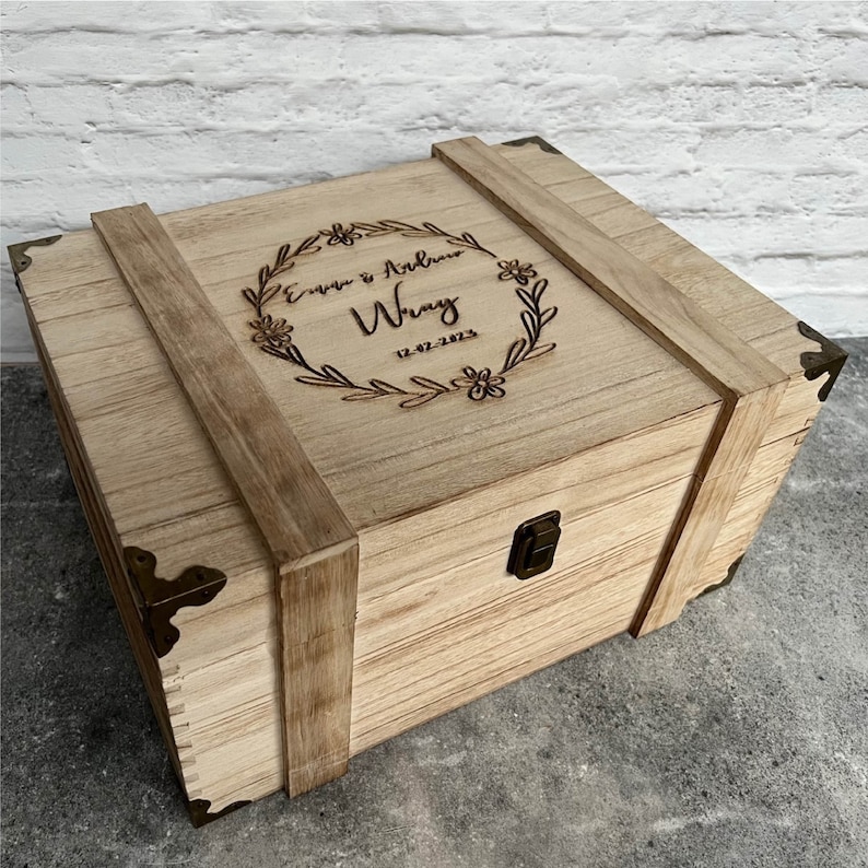 May include: A light-coloured wooden box with a hinged lid and metal clasp. The box is decorated with corner brackets and wooden straps. The lid features a floral wreath design with the names "Emma & Andrew Wray" and the date "12-02-2023" engraved in the wood. this can be changed to your text