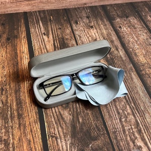May include: Black-framed eyeglasses inside a gray case with a matching microfiber cleaning cloth. The glasses have a rectangular shape and are resting on a wooden surface.