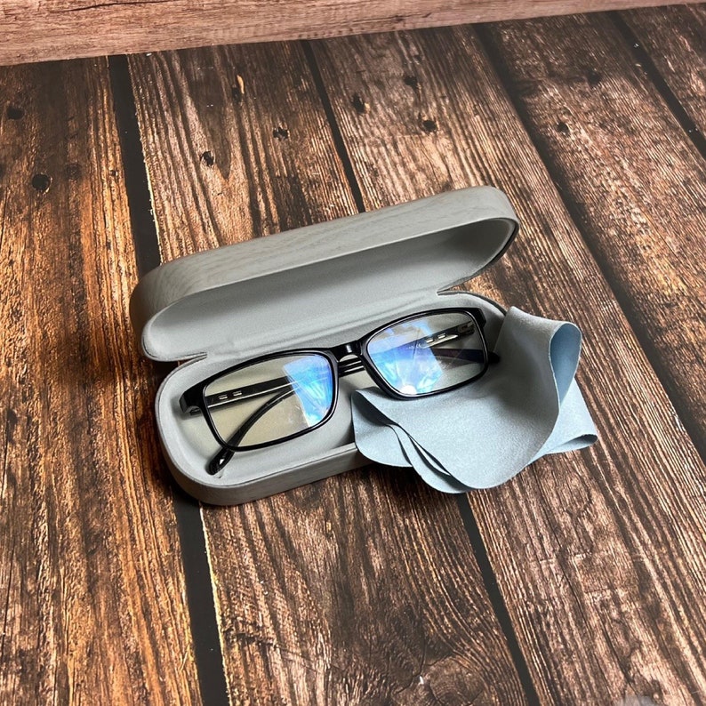 May include: Black-framed eyeglasses inside a gray case with a matching microfiber cleaning cloth. The glasses have a rectangular shape and are resting on a wooden surface.