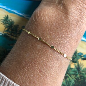 Puede incluir: Una delicada pulsera dorada con peque&ntilde;os abalorios redondos espaciados a lo largo de una fina cadena. La pulsera se muestra en una mu&ntilde;eca, con una manga de su&eacute;ter de color claro. El fondo incluye una escena tropical.