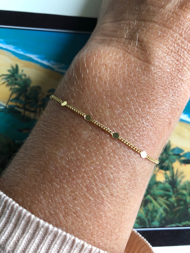 Puede incluir: Una delicada pulsera dorada con peque&ntilde;os abalorios redondos espaciados a lo largo de una fina cadena. La pulsera se muestra en una mu&ntilde;eca, con una manga de su&eacute;ter de color claro. El fondo incluye una escena tropical.