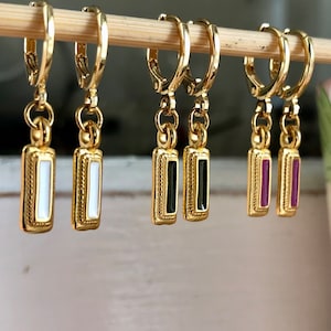 Peut inclure: Quatre paires de boucles d'oreilles créoles en or avec un charme rectangulaire suspendu. Les charmes sont blancs, noirs et deux nuances de violet.