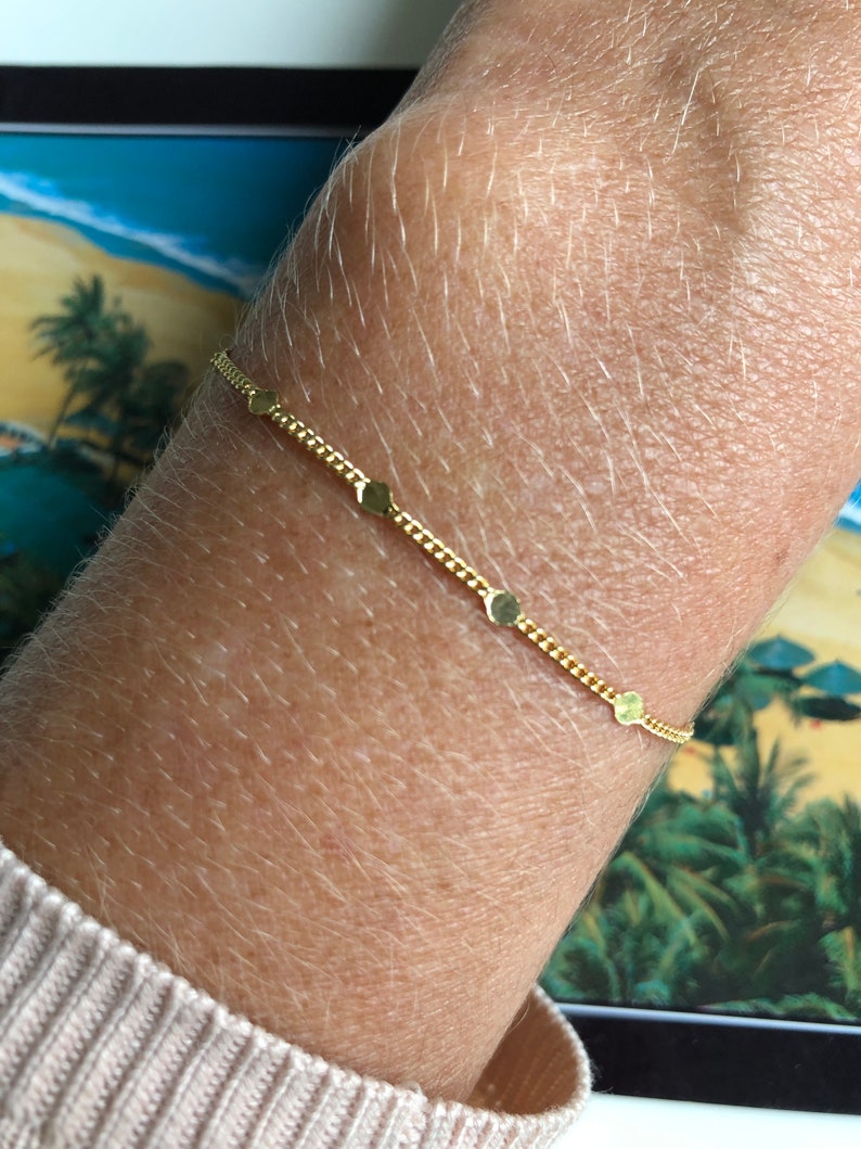 Puede incluir: Una delicada pulsera dorada con una cadena y peque&ntilde;os dijes planos en forma de coraz&oacute;n. La pulsera se lleva en la mu&ntilde;eca, mostrando su elegancia sencilla. El color dorado proporciona un c&aacute;lido contraste con la piel.