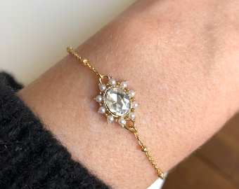 Puede incluir: Una pulsera dorada con un cristal transparente de forma ovalada en el centro, rodeado de pequeñas perlas blancas. El cristal es el punto focal, con las perlas dispuestas en forma de sol. La delicada cadena está conectada al diseño central.