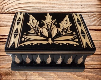 May include: A black wooden box with a decorative, intricate, carved design on the lid. The design is in a light wood colour and features a floral pattern. The box has a hinged lid and a secret compartment.