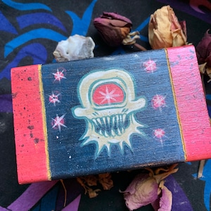 May include: A small wooden box with a red and gold border. The lid is painted with a black and white illustration of a skull with a single eye and a toothy grin. The background is a dark blue with white stars.