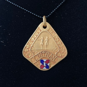 May include: A gold-colored pendant necklace with a diamond-shaped charm featuring two figures and the word "CENTENARIO". The charm also has a small red, white, and blue flag with a number "1" on it.