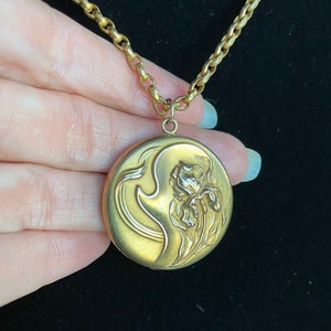 May include: A gold locket necklace with a chain. The locket is round and features an embossed design of an iris flower.