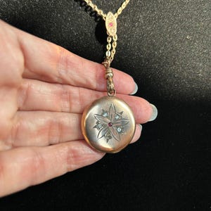 Pode incluir: Um colar dourado com um pingente redondo. O pingente apresenta um design floral com uma pedra vermelha central e pedras transparentes. A corrente é fina e o fundo é preto.