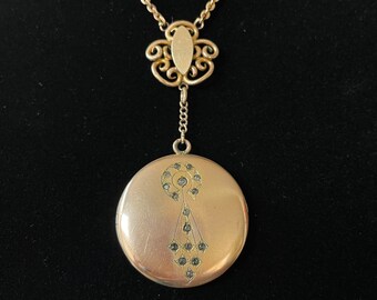 Può includere: Collana con medaglione dorato. Il pendente rotondo presenta un design geometrico con piccoli abbellimenti scuri. La catena e la chiusura sono decorative, in stile filigrana.