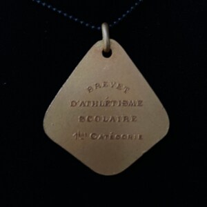 May include: A gold-colored metal pendant with a diamond shape. The pendant has the text "Brevet d'Athl&eacute;tisme Scolaire 1&egrave;re Cat&eacute;gorie" engraved on it.