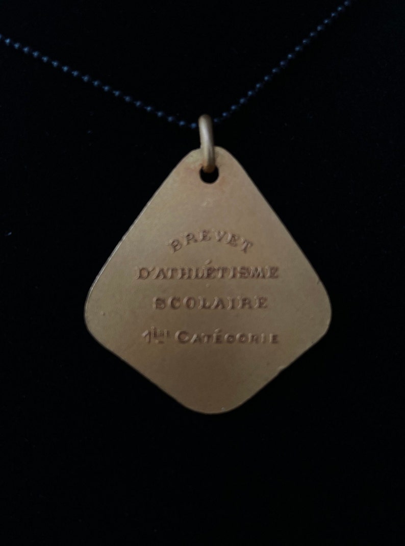 May include: A gold-colored metal pendant with a diamond shape. The pendant has the text "Brevet d'Athl&eacute;tisme Scolaire 1&egrave;re Cat&eacute;gorie" engraved on it.