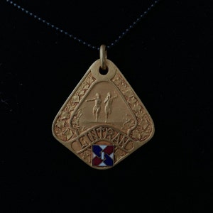May include: A gold-colored pendant necklace with a triangular charm featuring two figures running and the word "ENTRANCE" in a decorative font. The charm also has a small red, white, and blue square with the number "1" in the center.