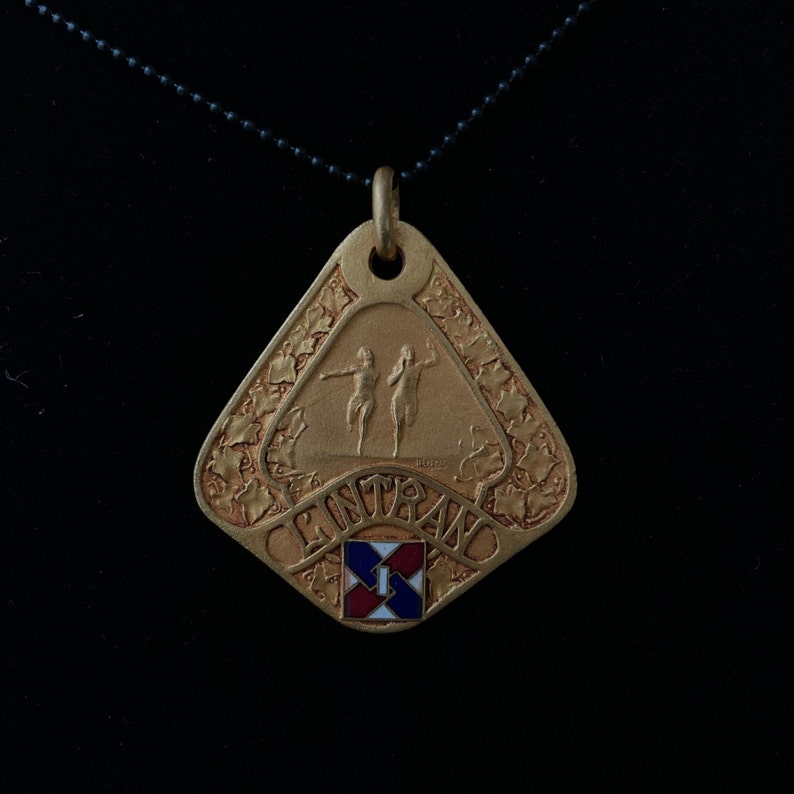 May include: A gold-colored pendant necklace with a triangular charm featuring two figures running and the word "ENTRANCE" in a decorative font. The charm also has a small red, white, and blue square with the number "1" in the center.