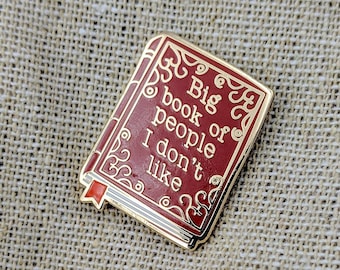 May include: A red enamel pin shaped like a book with gold trim. The pin says "Big book of people I don't like".