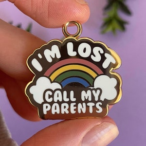 Puede incluir: Encanto de metal dorado con un arcoíris y el texto "I'm Lost Call My Parents".