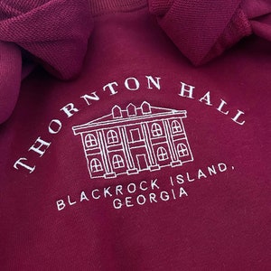 Puede incluir: Sudadera color burdeos con texto bordado en blanco que dice "Thornton Hall, Blackrock Island, Georgia". El texto está en forma de arco sobre una pequeña imagen bordada de un edificio.